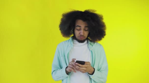 Egy afrikai frizurájú, narancssárga hátterű fiatalember ránéz a telefonra és boldog. Érzelmek egy színes háttér — Stock videók