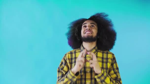 En ung man med en afrikansk frisyr på blå bakgrund korsar fingrarna och ber. Känslor på en färgad bakgrund. — Stockvideo