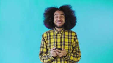 Mavi arka planda Afrikalı saç stili olan genç bir adam telefona bakar ve mutludur. Renkli bir arkaplanda duygular