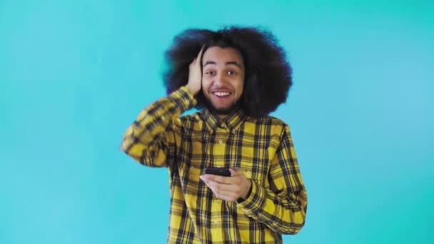 Un joven con un peinado africano sobre un fondo azul mira el teléfono y está felizmente sorprendido. Emociones sobre un fondo coloreado — Vídeos de Stock