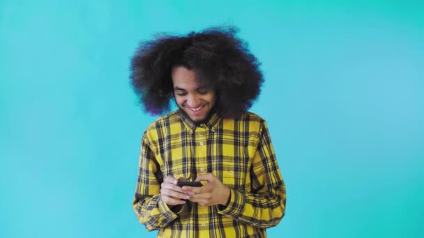 Молодой человек с африканской прической на синем фоне смотрит на телефон и счастливо пишет кому-то смс. Эмоции на цветном фоне — стоковое видео