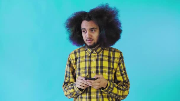 Młody mężczyzna z afrykańską fryzurą na niebieskim tle patrzy na telefon i ma wątpliwości. Emocje na kolorowym tle — Wideo stockowe