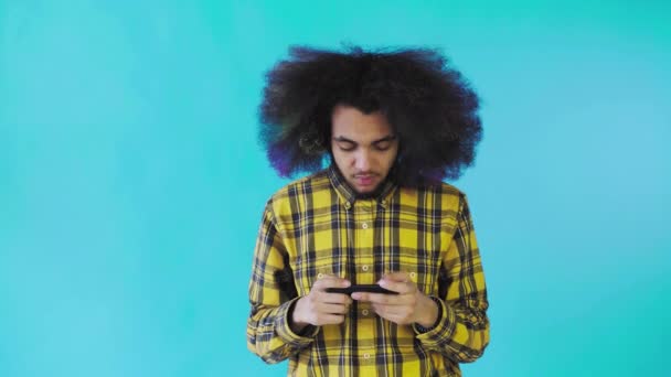 Um jovem com um penteado africano num fundo azul está a tocar ao telefone. Em um fundo colorido — Vídeo de Stock