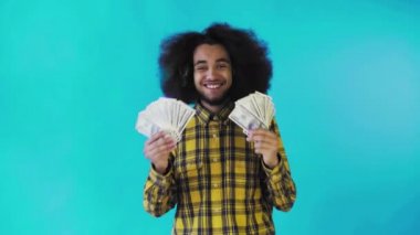 Elinde banknot yelpazesi olan mutlu bir Afro-Amerikalı adam mavi bir arka planda tek başına kameraya bakıyor.