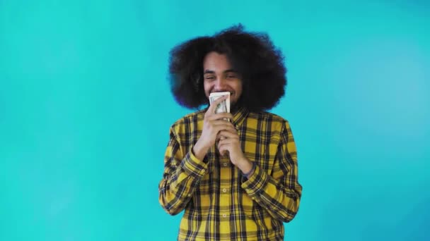 Egy boldog afro-amerikai férfi, aki bankjegyeket tart a kezében, megcsókolja és a kamerába néz, elszigetelve áll a kék háttérben. — Stock videók