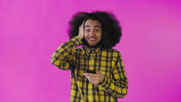 Um jovem com um penteado africano em um fundo rosa olha para o telefone e fica feliz surpreso. Emoções em um fundo colorido — Vídeo de Stock