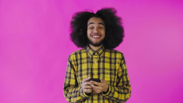 Un giovane con un'acconciatura africana su uno sfondo rosa guarda il telefono ed è felice. Emozioni su sfondo colorato — Video Stock