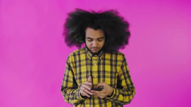 Молодой человек с африканской прической на розовом фоне вытаскивает свой телефон, но он не работает. Эмоции на цветном фоне — стоковое видео