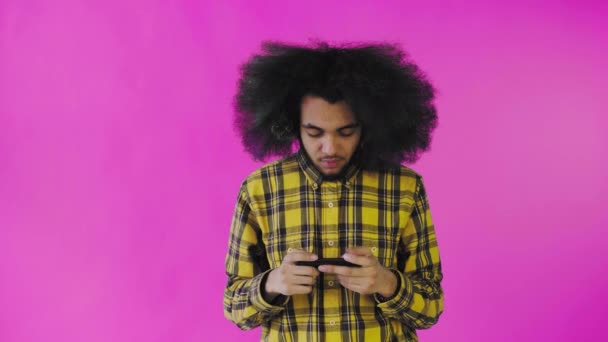 Un joven con un peinado africano sobre un fondo rosa está jugando por teléfono. Sobre un fondo de color — Vídeos de Stock