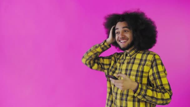 En ung mand med en afrikansk frisure på en lyserød baggrund ser på telefonen og er glad overrasket. Følelser på en farvet baggrund – Stock-video