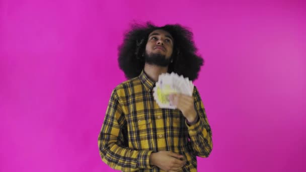 Um homem afro-americano feliz segurando um fã de notas em suas mãos e olhando para a câmera, de pé isolado em um fundo rosa — Vídeo de Stock