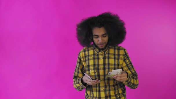 Un uomo afro-americano felice che tiene in mano le banconote, le conta e guarda la macchina fotografica, isolato su uno sfondo rosa — Video Stock
