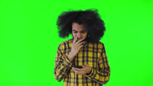 Een jonge man met een Afrikaans kapsel op een groene achtergrond kijkt naar de telefoon en is blij verrast. Emoties op een gekleurde achtergrond — Stockvideo