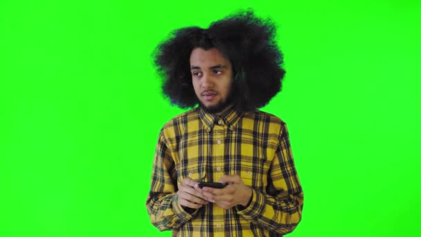 Un giovane con un'acconciatura africana su sfondo verde guarda il telefono e dubita. Emozioni su sfondo colorato — Video Stock