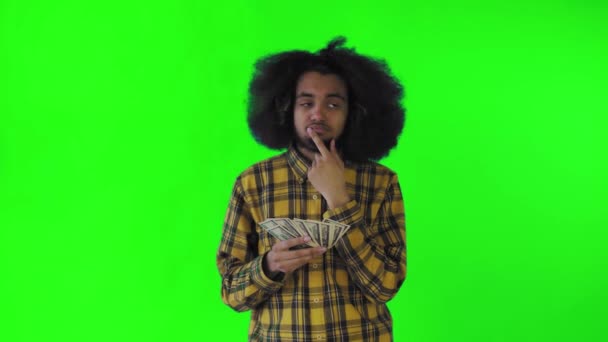 Un afro-americano feliz sosteniendo el dinero en sus manos y pensando en qué gastarlo, está aislado sobre un fondo verde — Vídeos de Stock