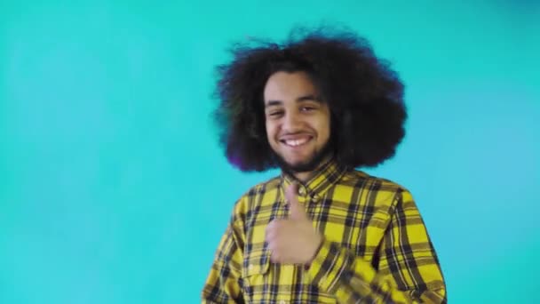 Un hombre afroamericano feliz mirando a la cámara, muestra un parecido con su mano, se encuentra aislado sobre un fondo azul — Vídeos de Stock