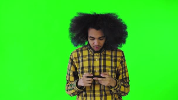 Un giovane con un'acconciatura africana su sfondo verde sta giocando al telefono. Su uno sfondo colorato — Video Stock