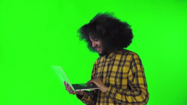 一个有着绿色背景的非洲发型的年轻人正在笔记本电脑上打字。在彩色背景上 — 图库视频影像