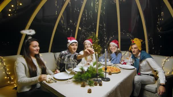 Friends celebrate New Year sitting at the dinner table. The young guy lights the candles. Students during a Christmas party at home. — Stockvideo