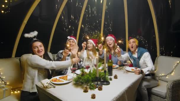 A group of friends are holding sparklers in their hands, smiling and looking at the camera. Friends celebrate the New Year at the dinner table. Students during a Christmas party at home. — Stockvideo