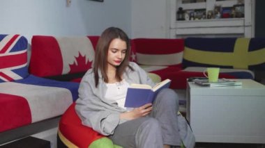 Genç bir kadın evinde bir sedirin üzerinde oturuyor ve kitap okuyor. Bir kadın boş zamanlarında rahat bir sedirin üzerinde dinleniyor. Hafta sonları iyi vakit geçir.