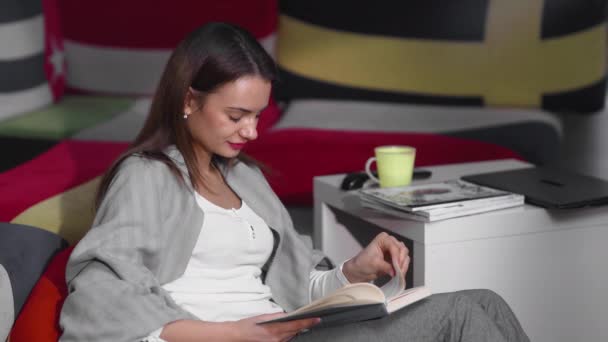 A young woman is sitting at home on an ottoman and reading a book. A woman is resting indoors on a comfortable ottoman in her free time. Have a nice free time on the weekend — Stock Video