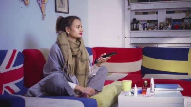 Side view en syg ung kvinde sidder på sofaen foran forskellige piller på bordet, ser tv – Stock-video