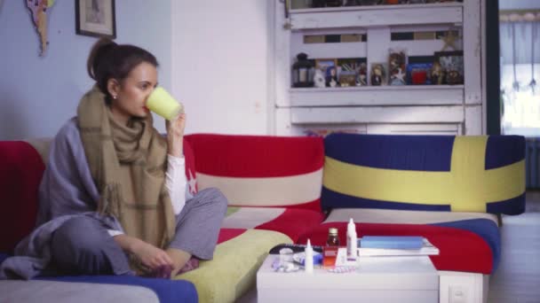 Vue de côté une jeune femme se sent mal et prend sa température. Une jeune femme assise sur le canapé, avec une écharpe, prend sa température. — Video