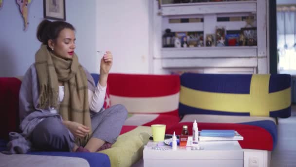 Side view a young woman feels unwell and takes her temperature. A young woman sitting on the sofa, with a scarf, takes her temperature. Drinks the medicine — Stock Video