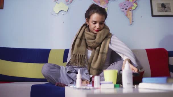 Vista lateral de una mujer joven enferma está sentado en el sofá delante de varias píldoras en la mesa, viendo la televisión — Vídeos de Stock