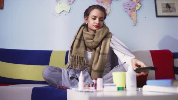 Een zieke jonge vrouw zit op de bank voor verschillende pillen op de tafel tv te kijken. — Stockvideo