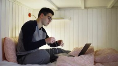 Yatakta genç bir adam oturuyor ve bir dizüstü bilgisayarla çalışıyor, kulaklıkları takıyor ve klavyeye yazıyor, geceleri çalışıyor, uzaktan kumandayla evden çalışıyor..