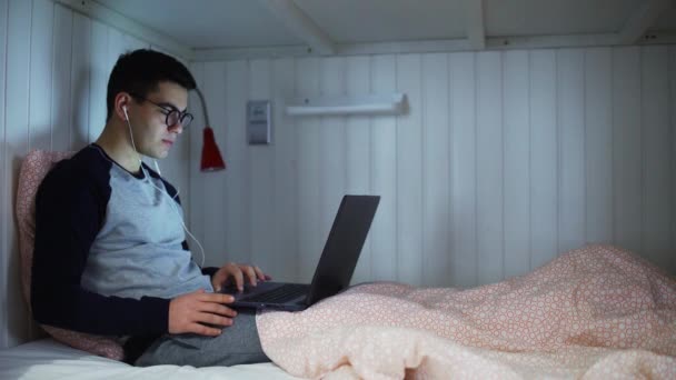 Vue de côté un jeune homme est allongé sur un lit et travaille sur un ordinateur portable, tapant sur un clavier, travaillant la nuit, travaillant à distance de la maison la nuit. — Video