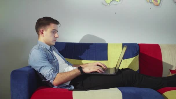 Vista lateral un hombre joven está acostado en el sofá y trabajando en un ordenador portátil, escribiendo en el teclado, trabajo remoto desde casa. — Vídeos de Stock