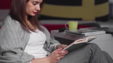 Genç bir kadın evinde bir sedirin üzerinde oturuyor ve kitap okuyor. Bir kadın boş zamanlarında rahat bir sedirin üzerinde dinleniyor. Hafta sonları iyi vakit geçir.