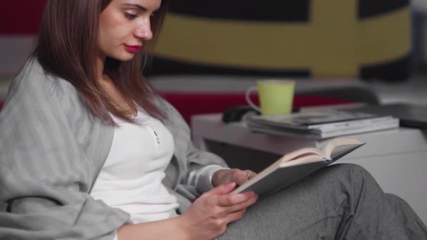 A young woman is sitting at home on an ottoman and reading a book. A woman is resting indoors on a comfortable ottoman in her free time. Have a nice free time on the weekend — Stock Video