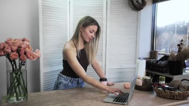 A young beautiful woman engaged in floristry, typing on a computer and drinking coffee. Flower shop. — Wideo stockowe