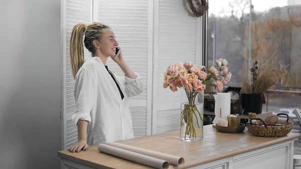 A young beautiful woman is engaged in floristry, talking on the phone. Flower shop. — Stockfoto