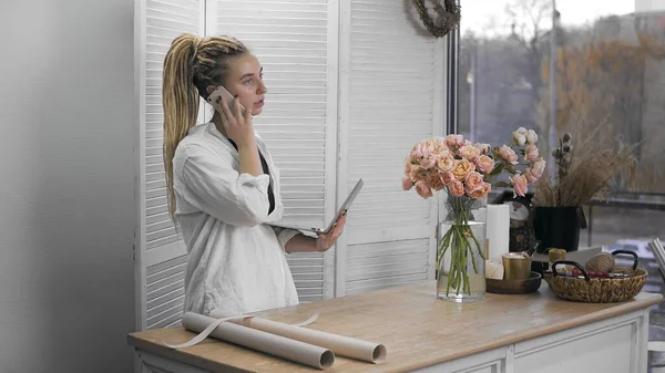 A young beautiful woman is engaged in floristry, talking on the phone and holding a computer in her hands. Flower shop. — Stockfoto