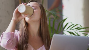Genç bir kadın ofiste bilgisayarın başında oturuyor ve kahve içiyor. İş kadını