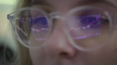 A close-up of a girl with glasses, her glasses reflect an Online Stock Market Chart Showing the Bivisand Bullish Trend of Bitcoin currency. In real time.