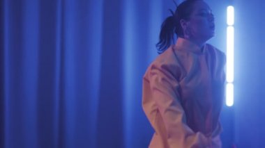 A beautiful girl is dancing indoors under blue lighting in a yellow suit. Dances