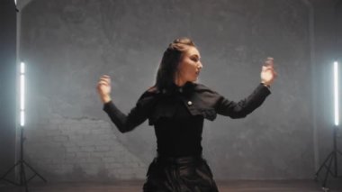 A beautiful girl is dancing indoors in a black suit. Dances
