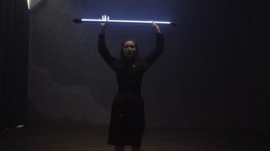 A beautiful girl is dancing indoors in a black suit with a blue lamp in her hands. Dances