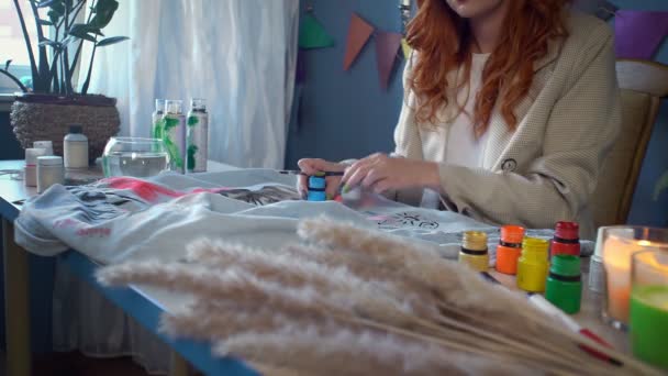 Close-up of a beautiful red-haired girl drawing with multicolored paints on clothes. Handmade work — Stockvideo