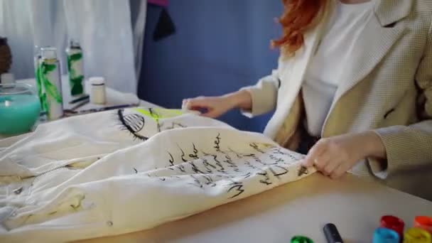 Close-up of a beautiful red-haired girl drawing with multicolored paints on clothes. Handmade work — Vídeo de Stock