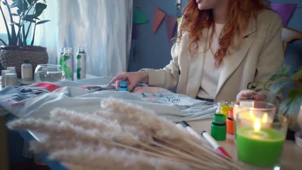 Close-up of a beautiful red-haired girl drawing with multicolored paints on clothes. Handmade work — Stock video
