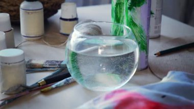 Close-up, the artist dilutes the paint with water, preparing to paint with paints. A womans hand lowers an artistic brush into the water, the blue paint dissolves.