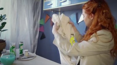 Güzel, kızıl saçlı bir kız elbiselerine renkli boyalar çiziyor. Kız bitirilmiş eserlere bakıyor. El yapımı iş