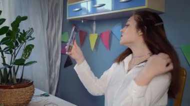 Beyaz gömlekli güzel bir kızın yakın çekimi boncuk süsleme oluşturur. Bir kız kendi elleriyle yapılan boncuklu bir mücevheri dener ve telefonuyla selfie çeker. El yapımı iş